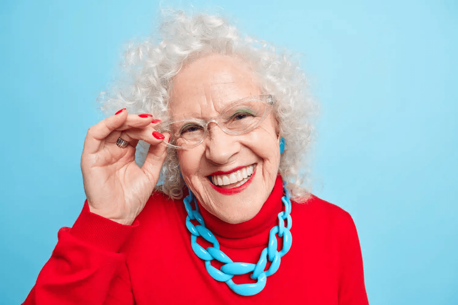 Elderly woman in white glasses wearing a red jumper and a big blue necklace smiles wide for the camera showing healthy teeth.
