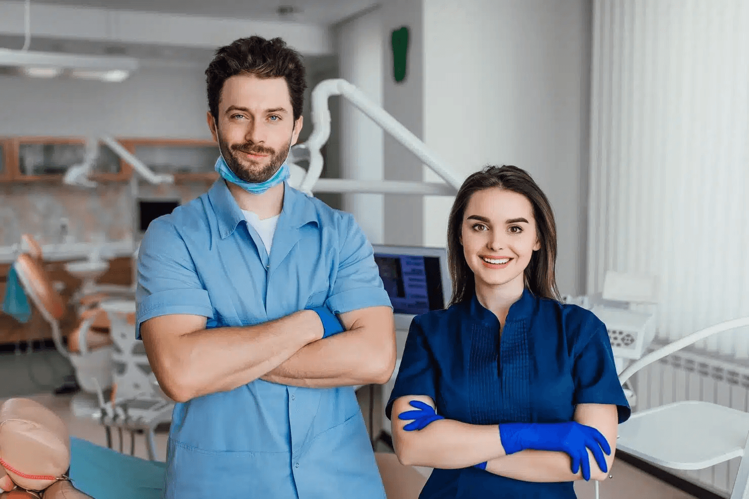 A male dentist and a female colleague smile for the camera with their arms crossed in a clinic setting.