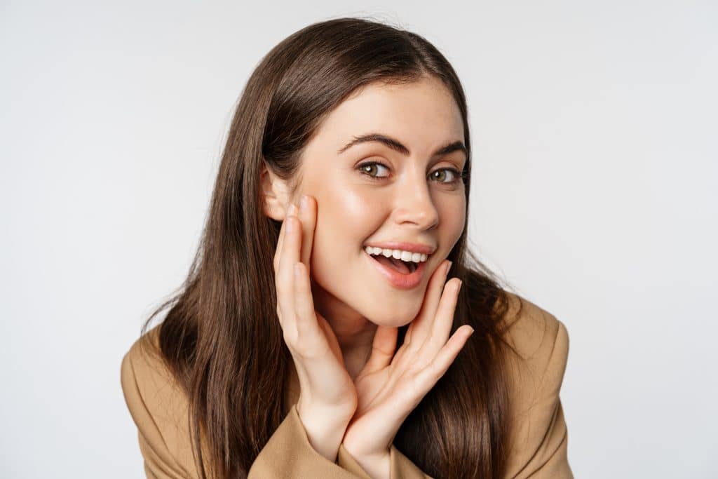 A close-up photograph of a happy woman examining her face.