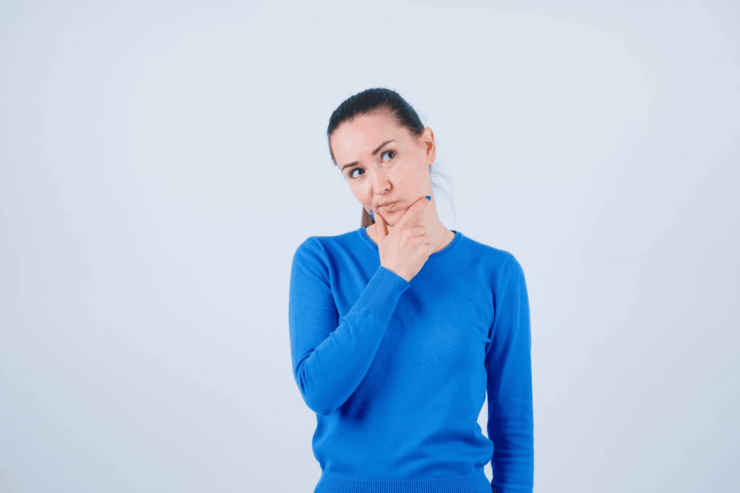 A woman in a bright blue top against a baby blue background thinks while holding her chin.
